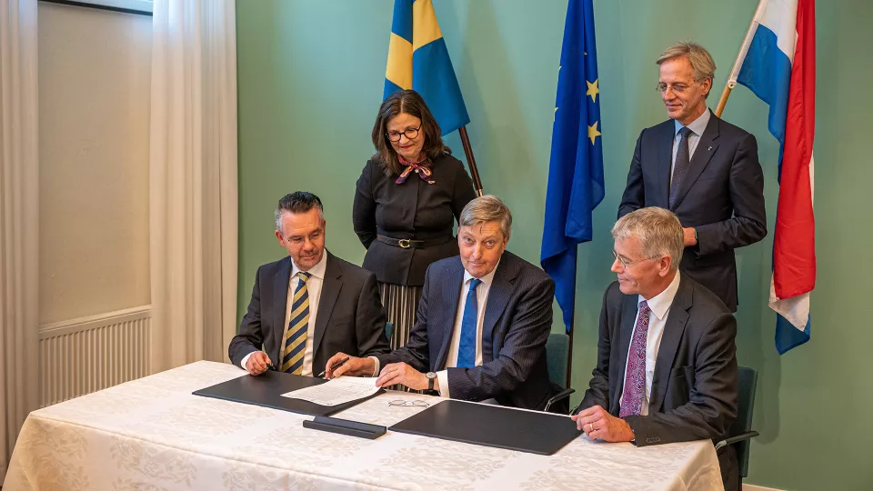 Memorandum being signed by three people with two people in the background, and the Swedish, Dutch and EU flags.
